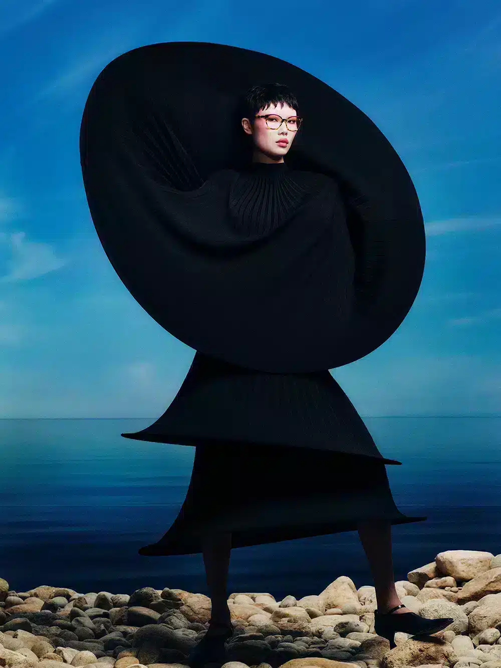 Femme portant des lunettes optiques de la collection SS25 d'Etnia Barcelona, avec une monture rouge et un style avant-gardiste, devant un paysage marin.