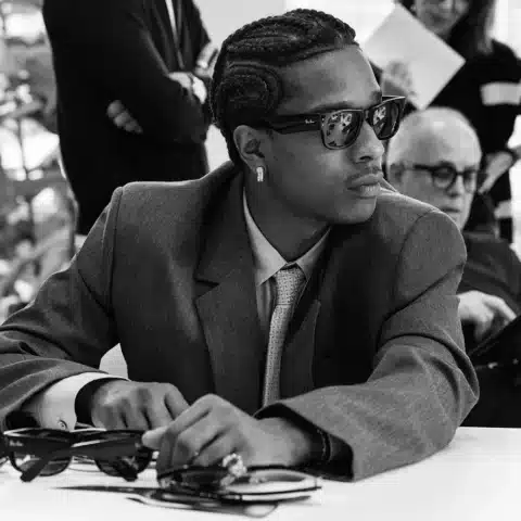 A$AP Rocky x Ray-Ban. A$AP Rocky en costume gris et lunettes Ray-Ban, observant attentivement une sélection de montures lors d’une réunion créative.